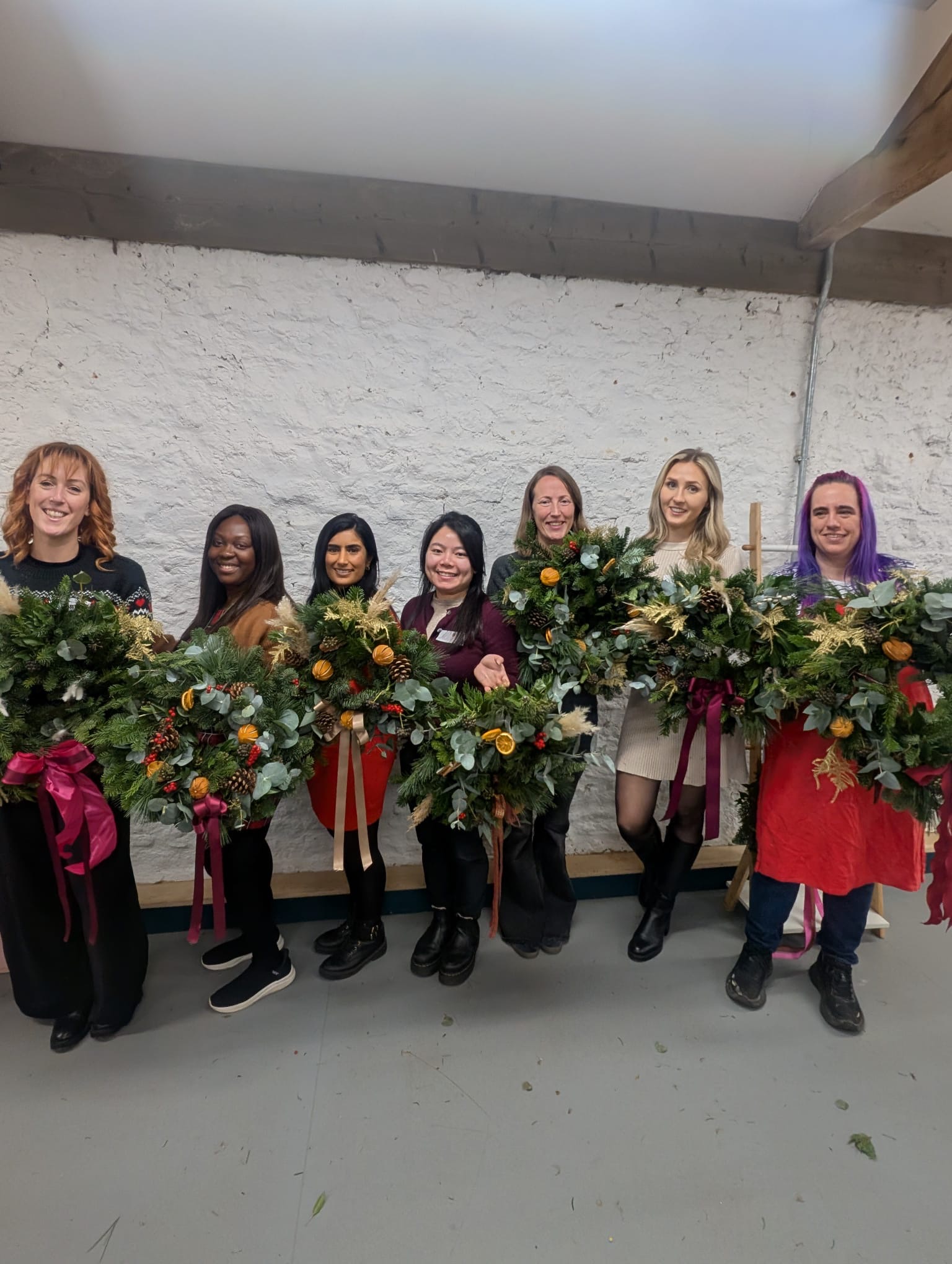 Seven women holding handmade Christmas wreaths decorated with foliage, dried oranges, pine cones and ribbons at a F.IN.A Connect wreath-making event.
